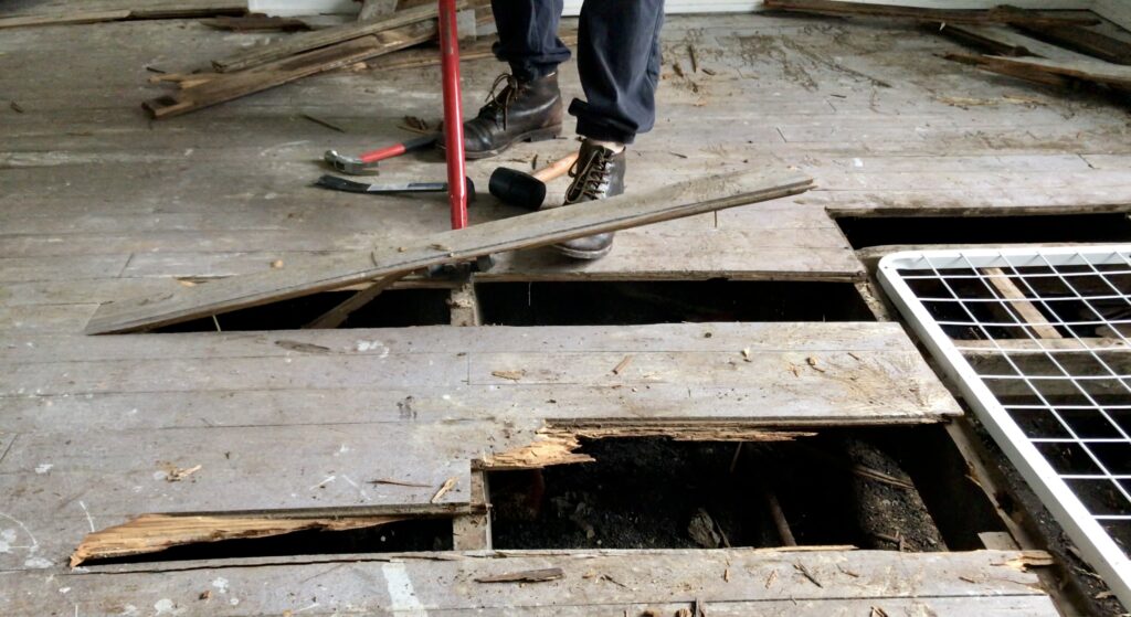 man using a pry bar to pull up wood flooring and a wood piece in the air being removed