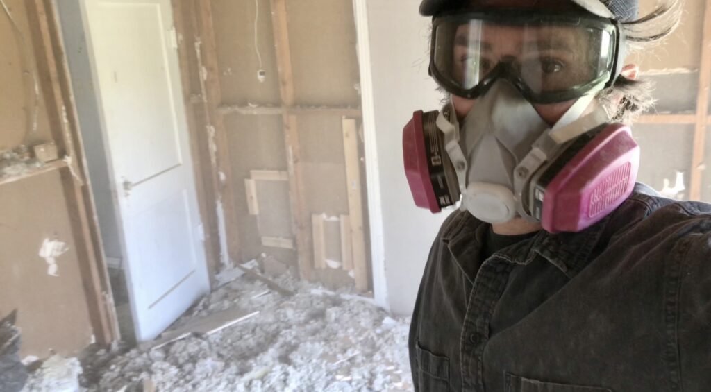 man with protective goggles and mask on with drywall and insulation pieces all around him with walls exposed in fixer upper