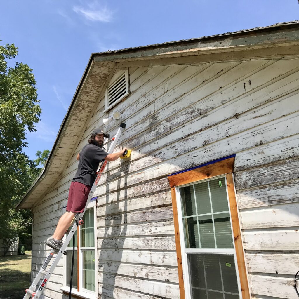 man on ladder scrubbing old original siding on little cottage
